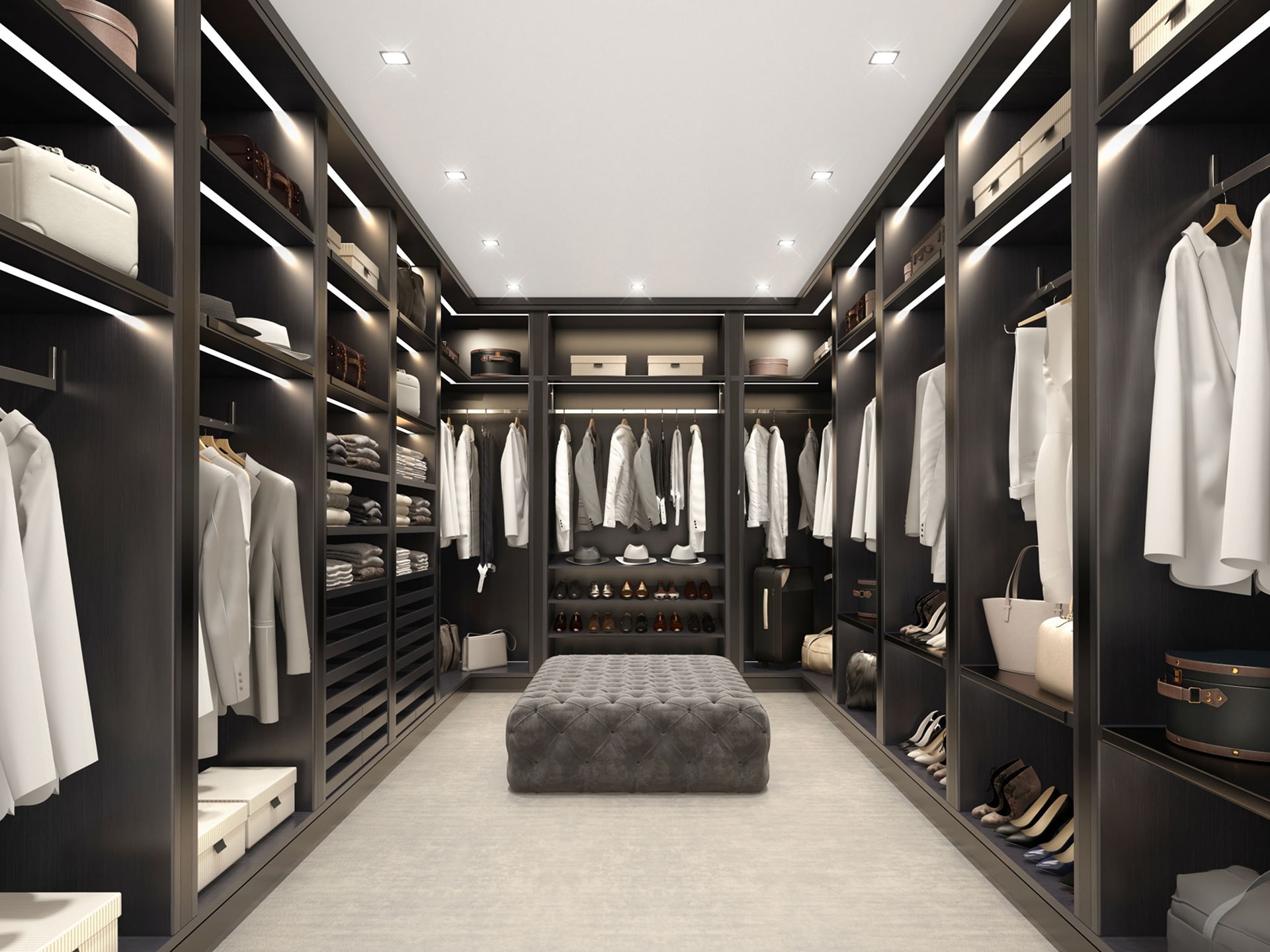 A luxurious and spacious fitted walk-in wardrobe boasts dark wood shelves and racks. Clothes, shoes, and accessories are neatly organized. An upholstered ottoman stands in the center of the room, which is brightly lit with recessed ceiling lights.