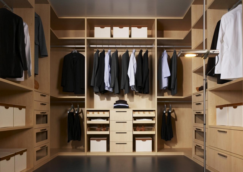 A spacious walk-in closet with built-in wardrobes, featuring neatly organized wooden shelves and drawers. The left and right sections display hanging clothes, mostly suits and shirts. The middle section features drawers and neatly folded clothes. Storage boxes are placed on various shelves.