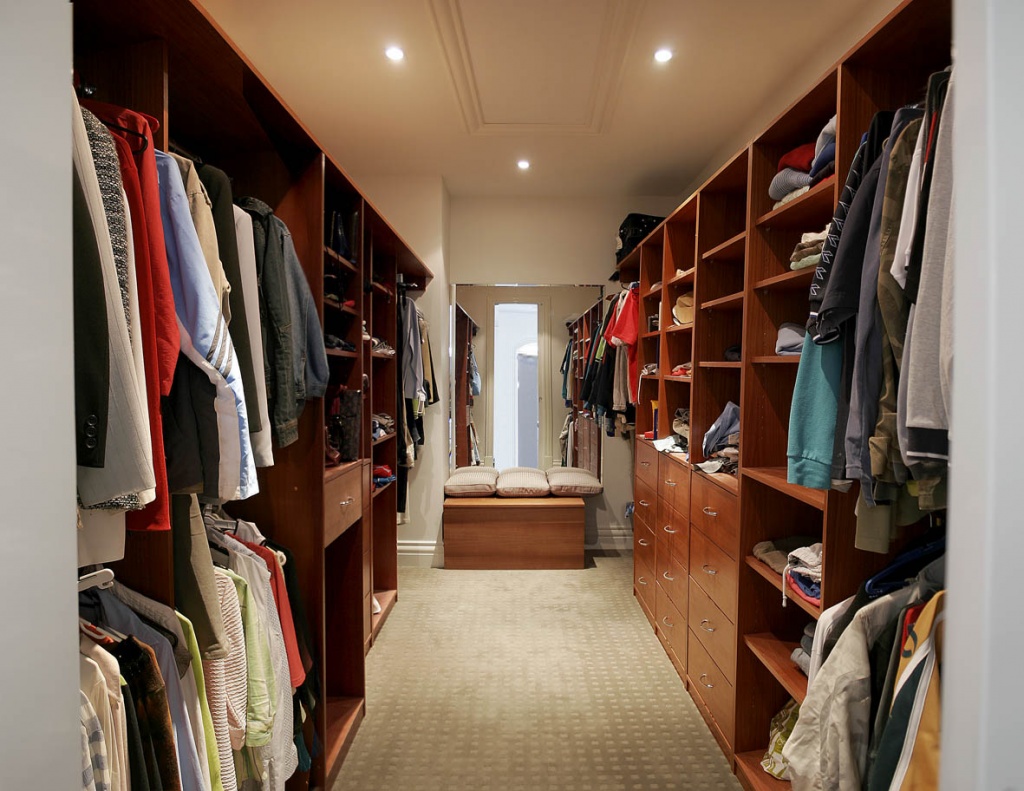 A spacious walk-in closet with built-in wardrobes, featuring neatly organized wooden shelves and drawers. The left and right sections display hanging clothes, mostly suits and shirts. The middle section features drawers and neatly folded clothes. Storage boxes are placed on various shelves.
