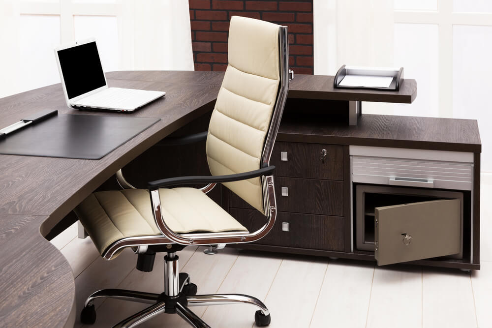A modern office with a dark wooden desk featuring bespoke joinery and a curved design with drawers. There is a beige cushioned office chair pushed in. The desk holds a laptop, a black desk pad, and some documents. An open safe is also visible in the bottom right of the image.