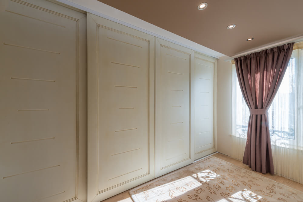 A room with beige walls features large built-in cabinets showcasing bespoke joinery with subtle horizontal indentations. The floor is carpeted in a matching beige tone with a floral pattern. There's a window with sunlight streaming in through brown curtains tied back with a matching tie.