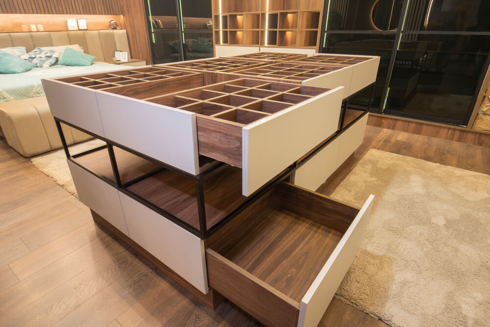 A modern, spacious walk-in closet with dark wood flooring features a central island with multiple drawers and open cubbies for storage. The room includes bespoke joinery, a comfortable beige armchair, a large mirrored wall, and ample lighting, creating a luxurious and organized space.