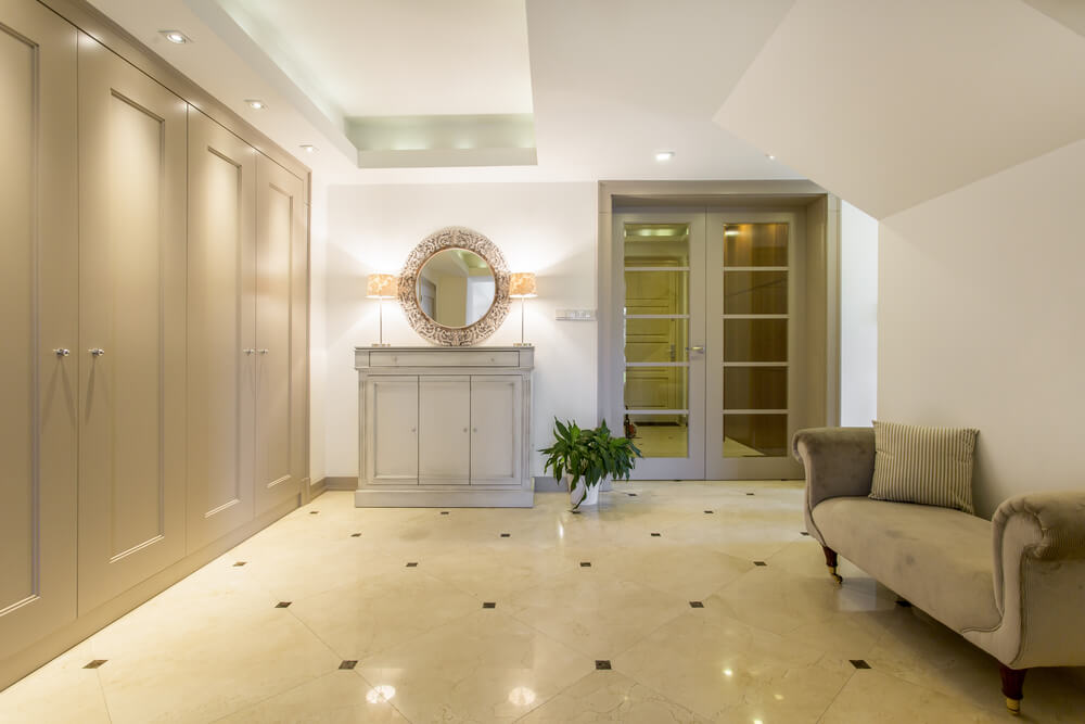 A cozy, well-lit hallway with beige walls and tile floors features bespoke joinery in the form of a small console table with a round mirror above it. The hallway includes a potted plant, a small sofa with a cushion on the right, and double doors with glass panels leading to another room.2