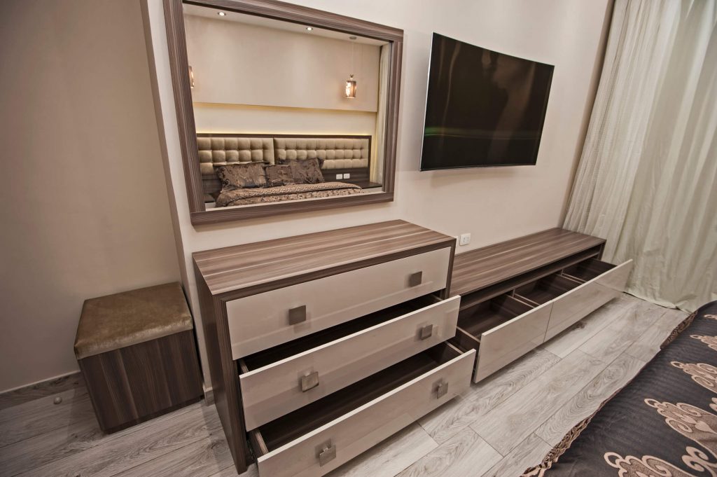 A modern bedroom with light wood flooring features a dresser with several drawers open, showcasing bespoke joinery. Above the dresser is a large, framed mirror reflecting the bed and two pendant lights. Next to the dresser is a wall-mounted flat-screen TV and a small side table.