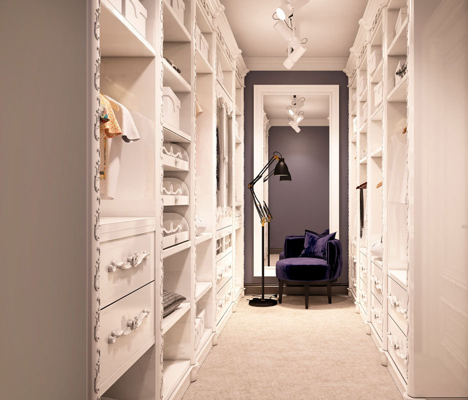 A well-organized walk-in closet with white shelves filled with clothes and accessories on both sides. At the end of the room, there's a large mirror reflecting a purple accent wall. Sliding door wardrobes add a touch of elegance while a dark chair with a floor lamp sits in front of the mirror.