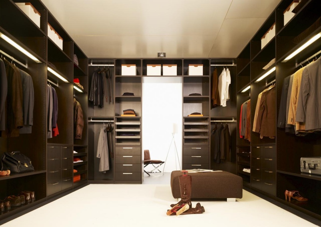 A spacious walk-in wardrobe features dark wooden shelves and hanging racks filled with neatly organized clothing. An ottoman is centered on the light floor, with a pair of boots and shoes nearby. The closet extends to another room with a chair and floor lamp visible.