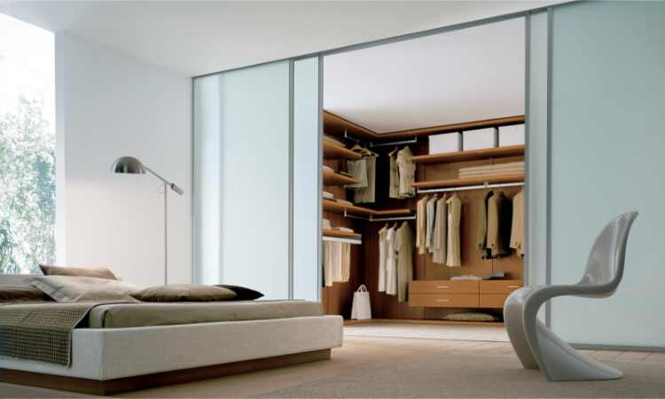 A spacious modern bedroom with a white bed, beige bedding, and a sleek white chair. In the background, an open closet with wooden shelves, hanging clothes, and drawers looks almost like luxurious walk-in wardrobes. Natural light illuminates the room through large windows with trees visible outside.