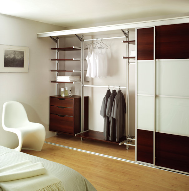 A modern, organized closet with an open design. Clothes hang on racks, and there are wooden shelves and a small drawer unit for storage. Sliding glass and wood doors are partially opened. A white chair and a bed with folded linens are visible nearby, resembling luxurious walk-in wardrobes.