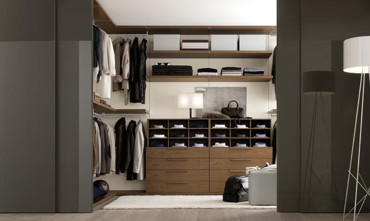 A modern, organized walk-in wardrobe with open shelving and drawers. Clothes are neatly hung and folded, with storage boxes on upper shelves. A light fixture and a framed picture adorn the space. A rug lies on the floor beside an armchair and a lamp.