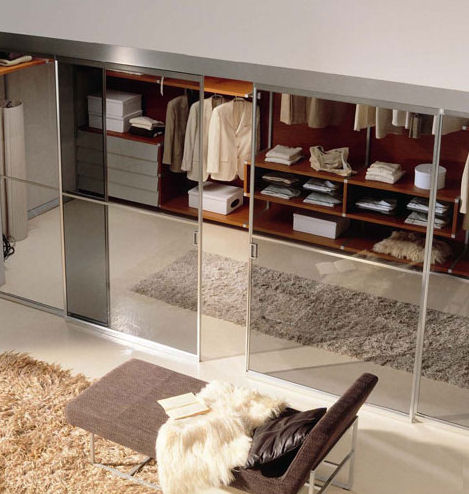 A modern closet with sliding glass doors reveals neatly organized clothing and shelves inside. The room has a beige carpet, a brown bench with a cushion, and a plush golden-brown rug. The clean, minimalist design of the space gives it a contemporary feel, reminiscent of luxurious walk-in wardrobes.