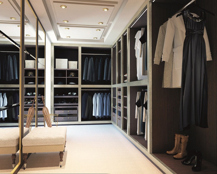 A spacious walk-in closet with neatly organized clothes on hangers and shelves, enhanced by built-in wardrobes. There is a mix of shirts, jackets, dresses, and trousers. The room has a bench in the center, a large mirror on one side, and recessed lighting in the ceiling. A pair of boots is on the floor.