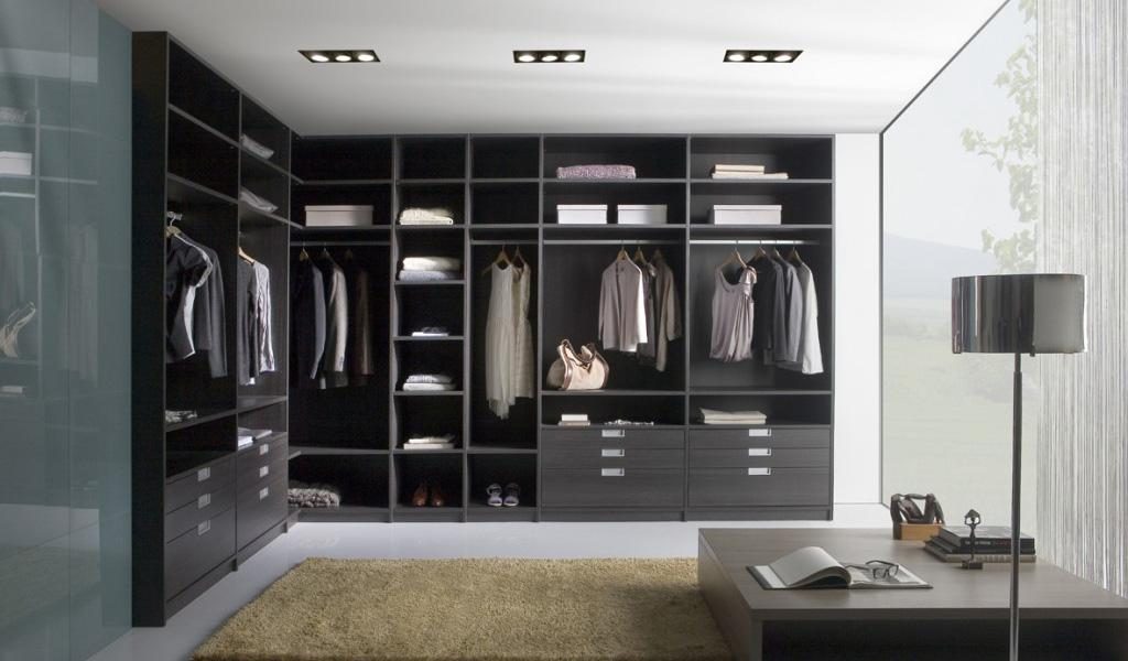 A modern walk-in wardrobe with dark shelving and drawers features clothing neatly hanging and folded under soft overhead lighting, complemented by a rug on the floor. A floor lamp and a sleek wooden bench with books add to the stylish, organized space while large windows allow natural light in.