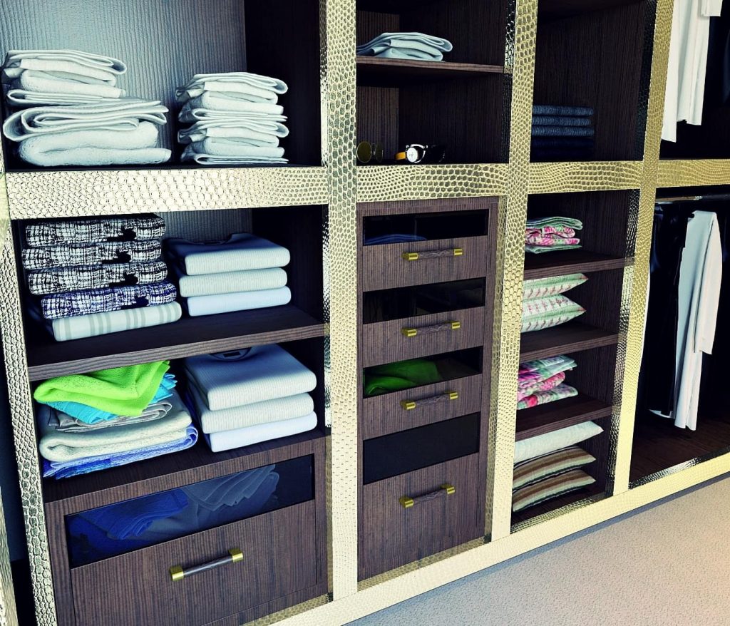 A neatly organized walk-in wardrobe with shelves holding folded towels, clothes, and linens. Four drawers are included in the center, and the structure features a gold metallic frame with dark wooden compartments. Hanging clothes are partially visible on the right side.