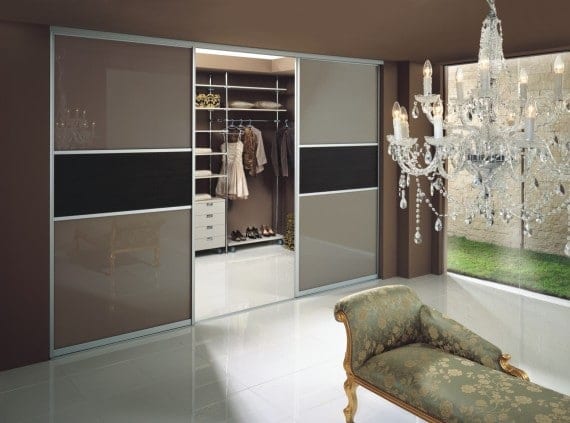 A modern beige walk-in wardrobe with sliding glass doors reveals shelves filled with clothes and shoes. An elegant chandelier hangs from the ceiling, and a luxurious upholstered chaise lounge sits in the foreground. A large window offers a view of a garden.