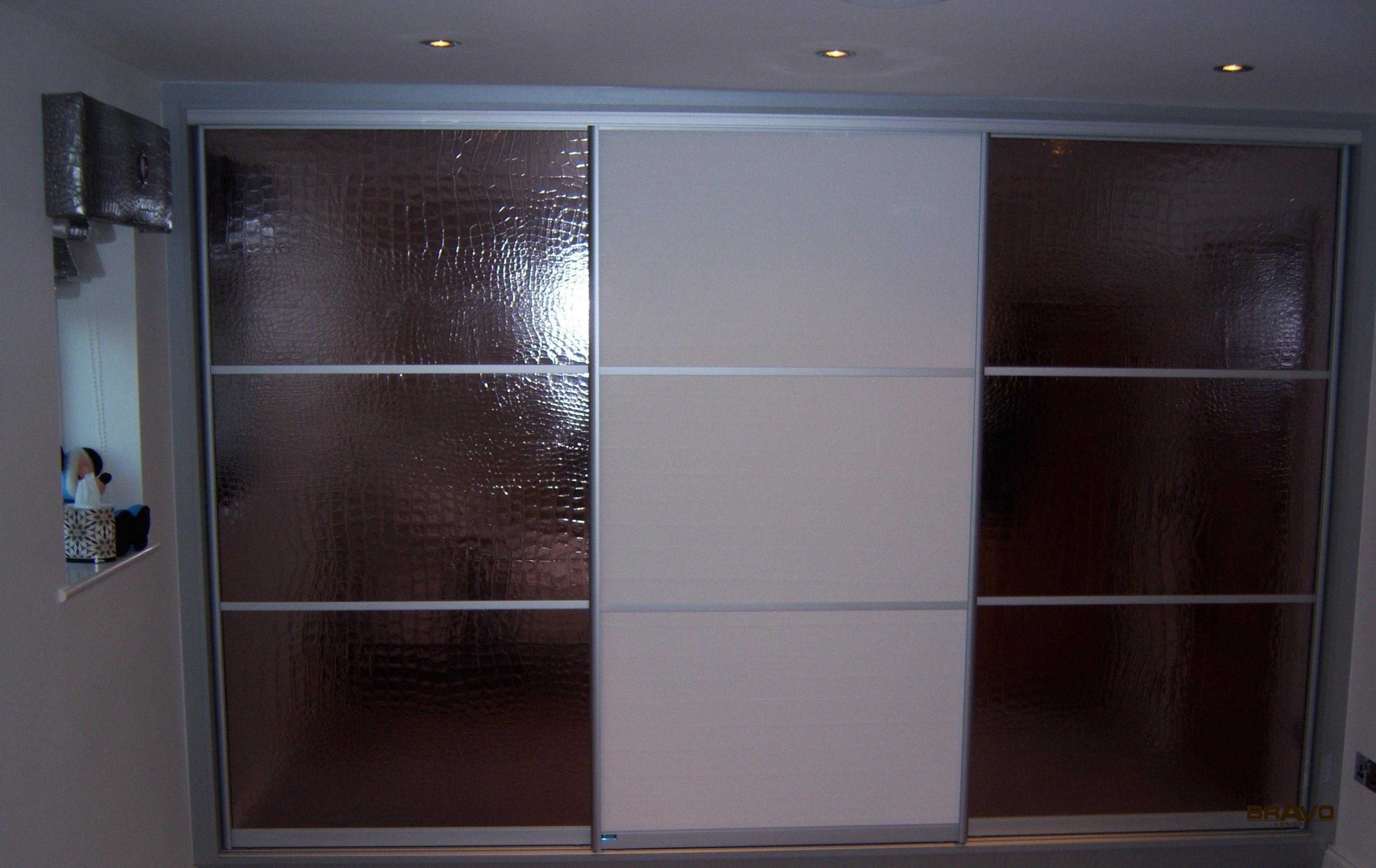 A large wardrobe with sliding doors, featuring textured glass panels and a combination of transparent and opaque sections, installed in a fitted bedroom with dim lighting.
