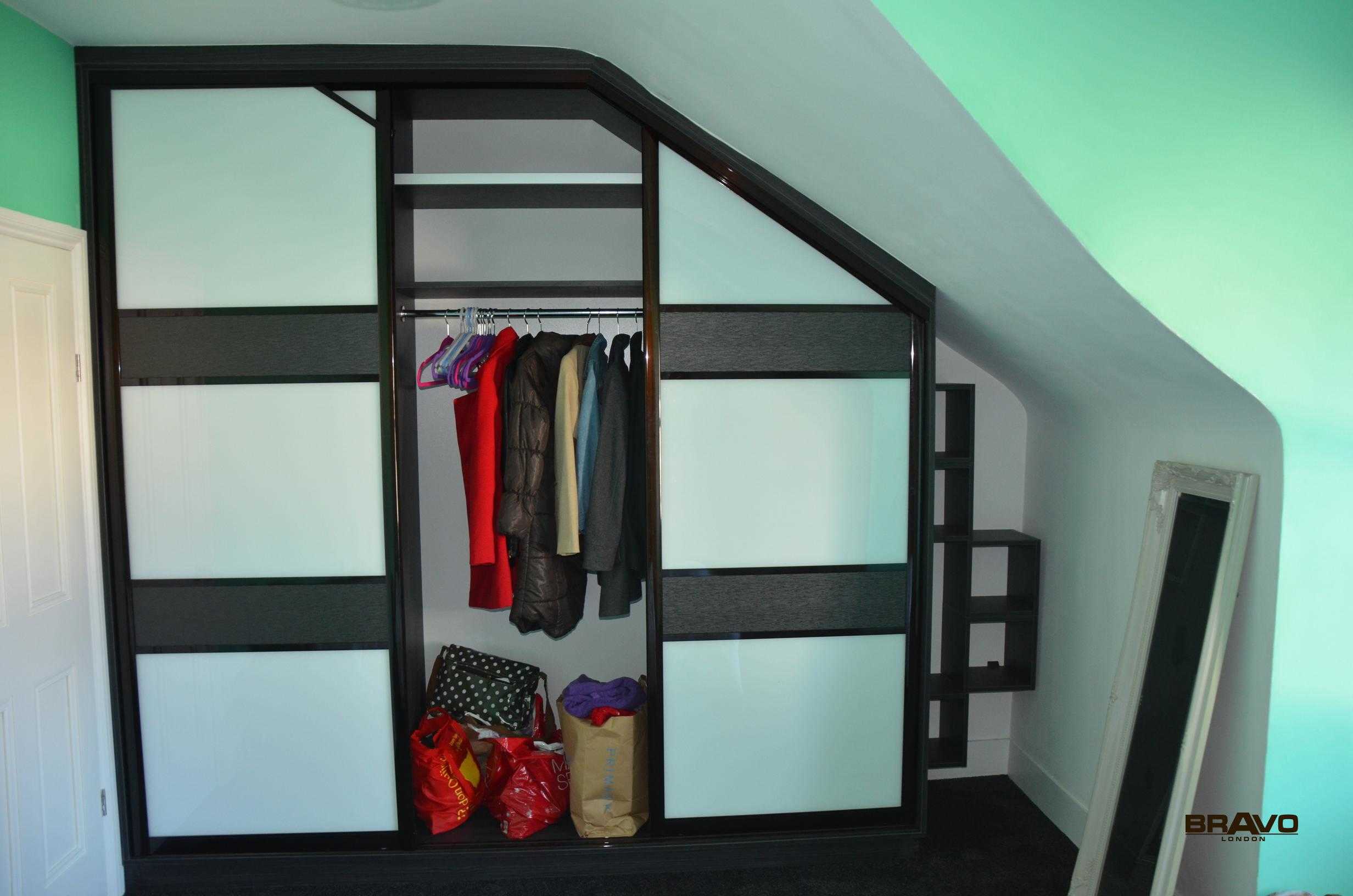 A large black-framed fitted wardrobe is installed into a slanted ceiling space, with partially open sliding doors revealing clothes and bags inside. The room has a green wall and wooden flooring.