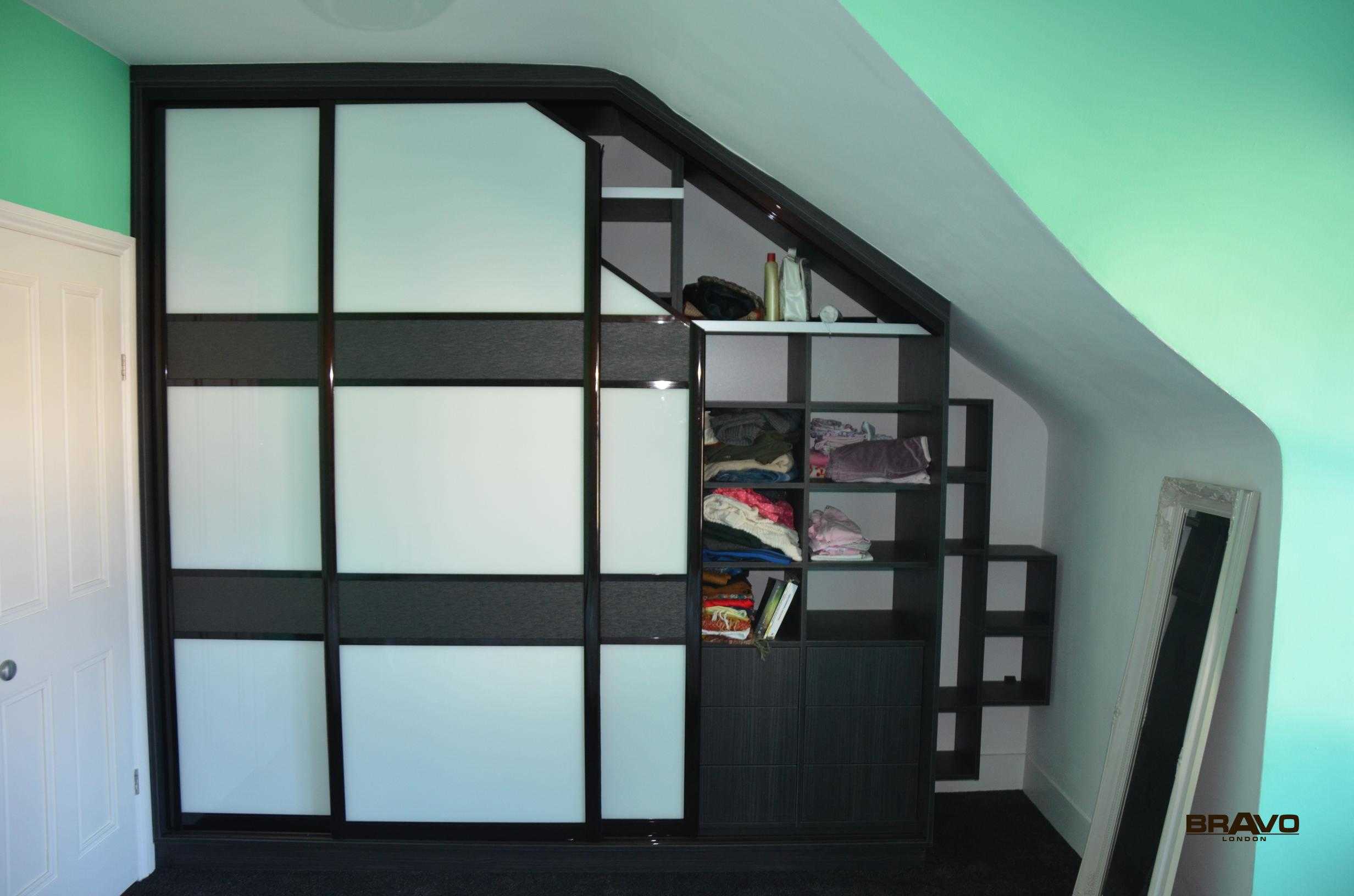 A modern walk-in wardrobe with black frames and frosted glass panels, built under a slanted ceiling in a room with mint-green walls.