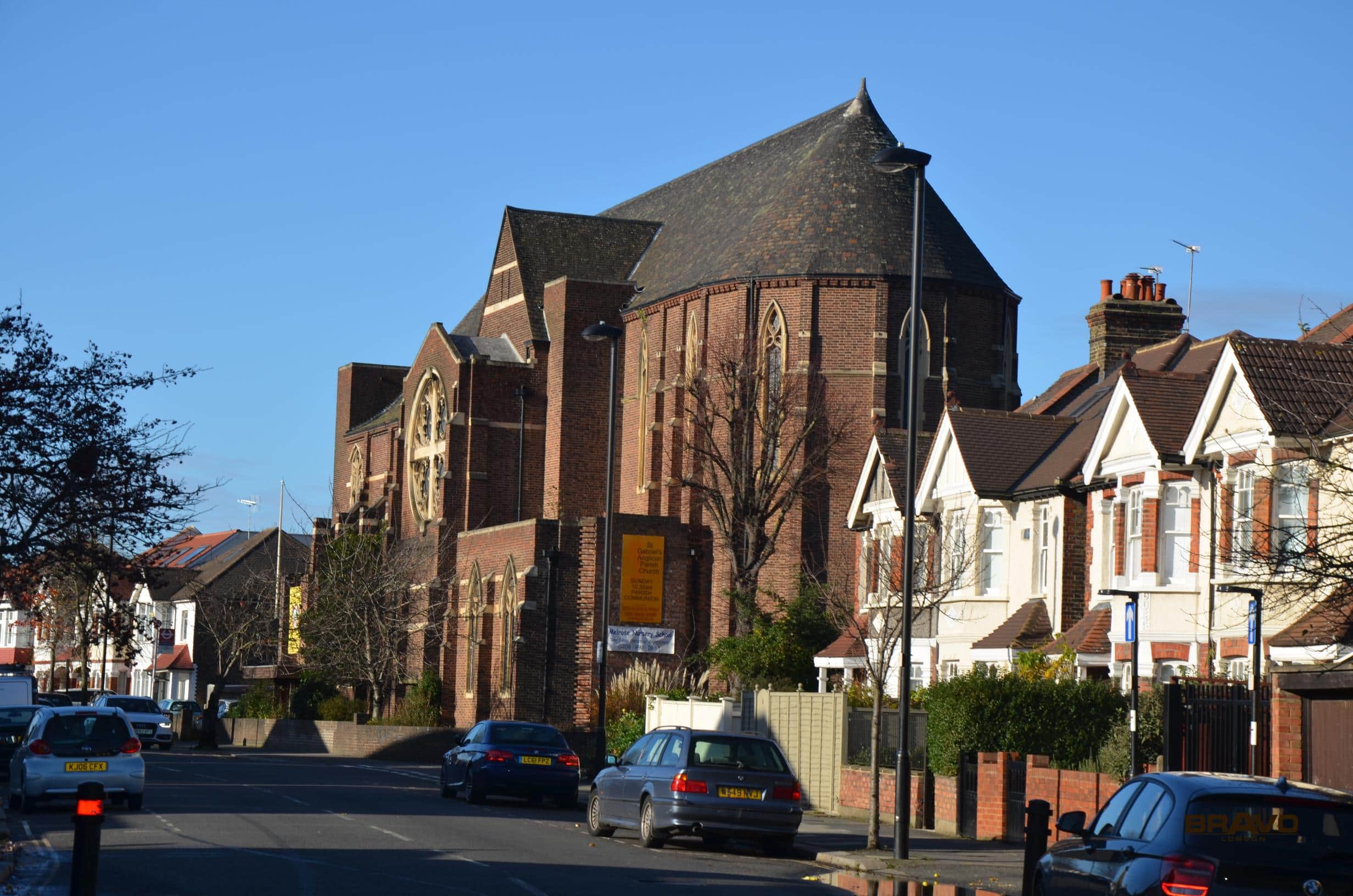 A large red brick church with a prominent arched roofline flanked by smaller buildings, featuring bespoke furniture, located on a sunny suburban street lined with parked cars.
