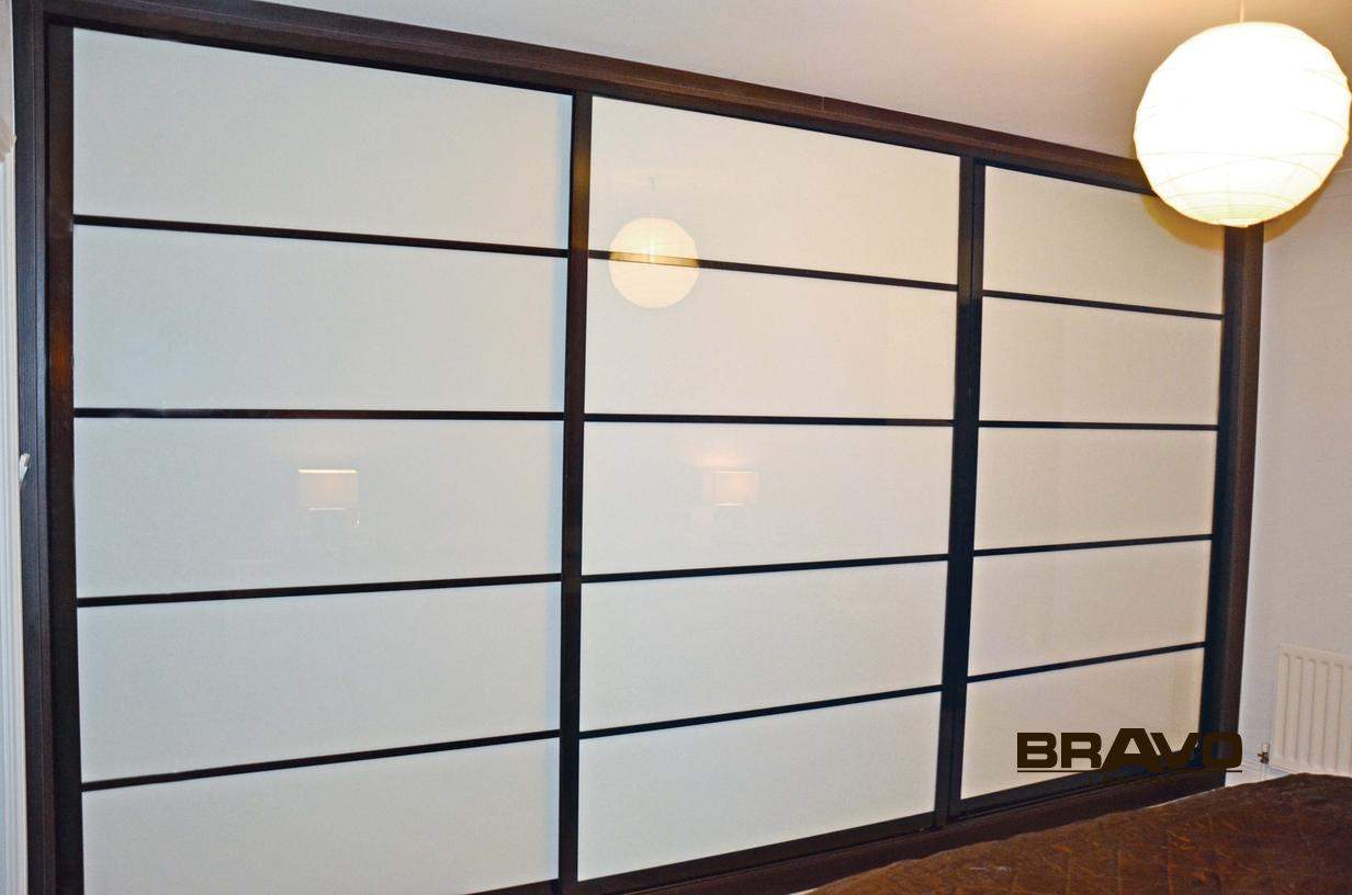 A large, modern sliding door wardrobe with frosted glass panels and black frames, installed in a room with beige walls, featuring a glimpse of a hanging light fixture.