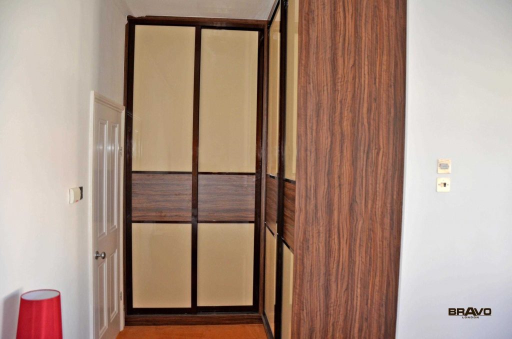 An indoor space featuring a large, dark wood fitted wardrobe with closed frosted glass doors, positioned adjacent to an open door leading to another room. A white wall and light switch are visible on the right.