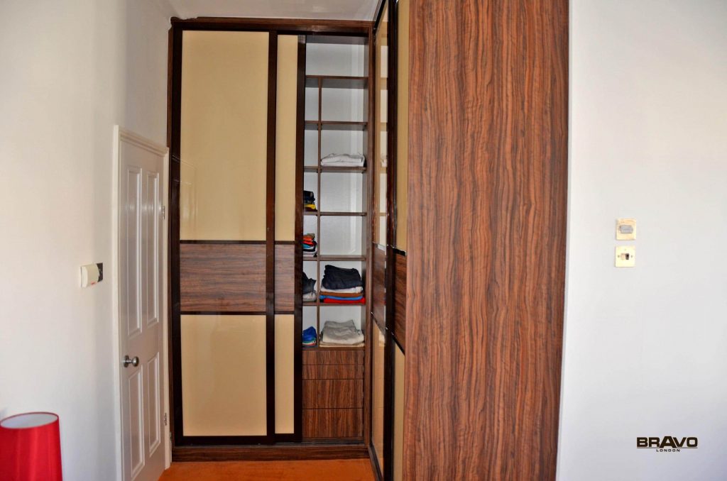 An open wooden wardrobe with sliding doors, shelves partially filled with neatly folded clothes and towels, viewed from the side with a glimpse of a room door in the background.
