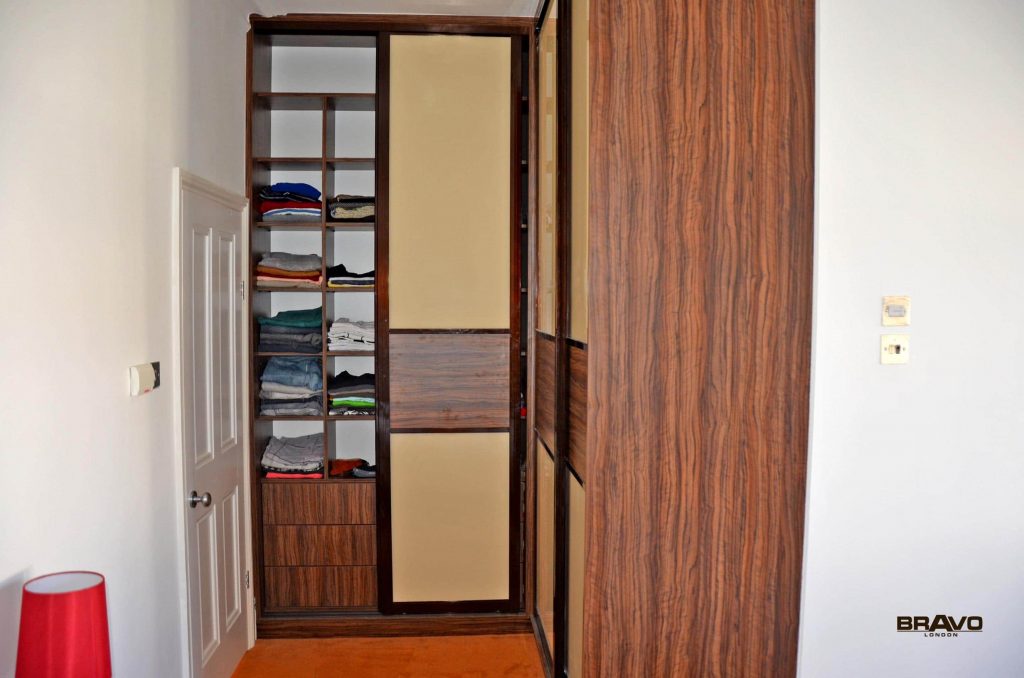 A partially open wooden wardrobe built into a wall, filled with neatly stacked clothes on shelves, in a room with a white wall and a red lamp on a cabinet, features bespoke furniture design ideal for fitted bedrooms.