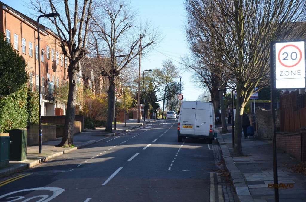 A quiet street on a clear day, lined with trees and buildings featuring fitted wardrobes, includes a delivery van driving down the road and a 20 mph speed limit sign.