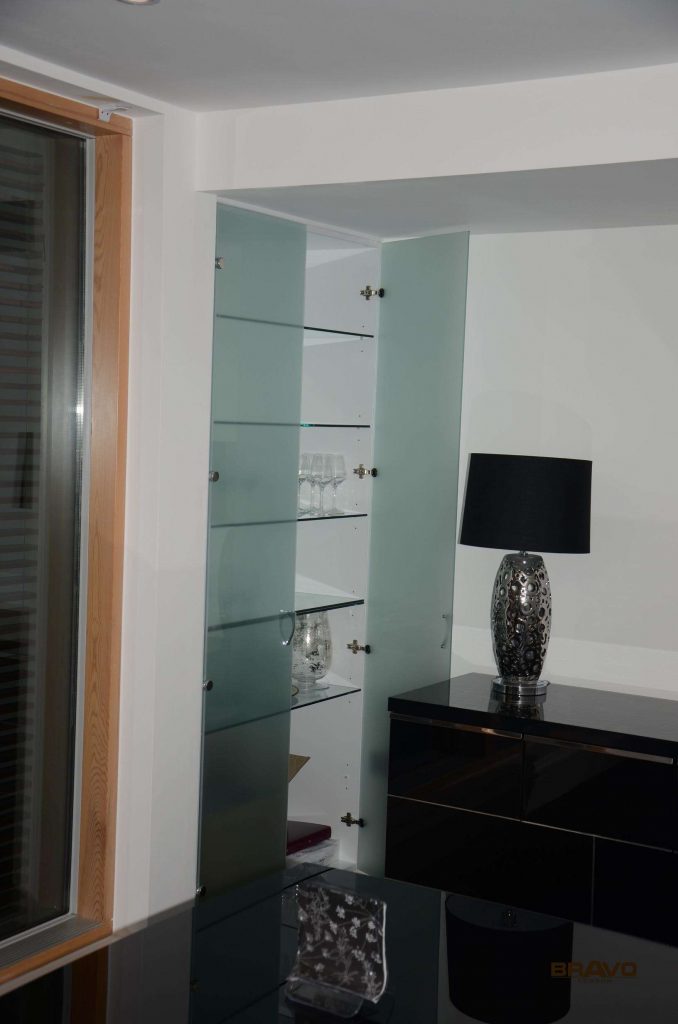 A modern interior featuring a glass display cabinet next to a window, and a black glossy fitted wardrobe with an ornate silver and black vase on top, situated in a room with glossy tiled flooring.