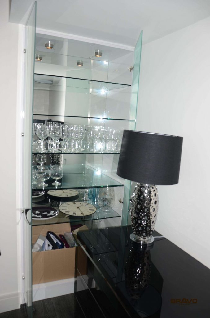 A sleek, modern display cabinet with glass shelves, stocked with various glasses, stands next to a black console table with a stylish lamp and decorative items in a fitted bedroom.