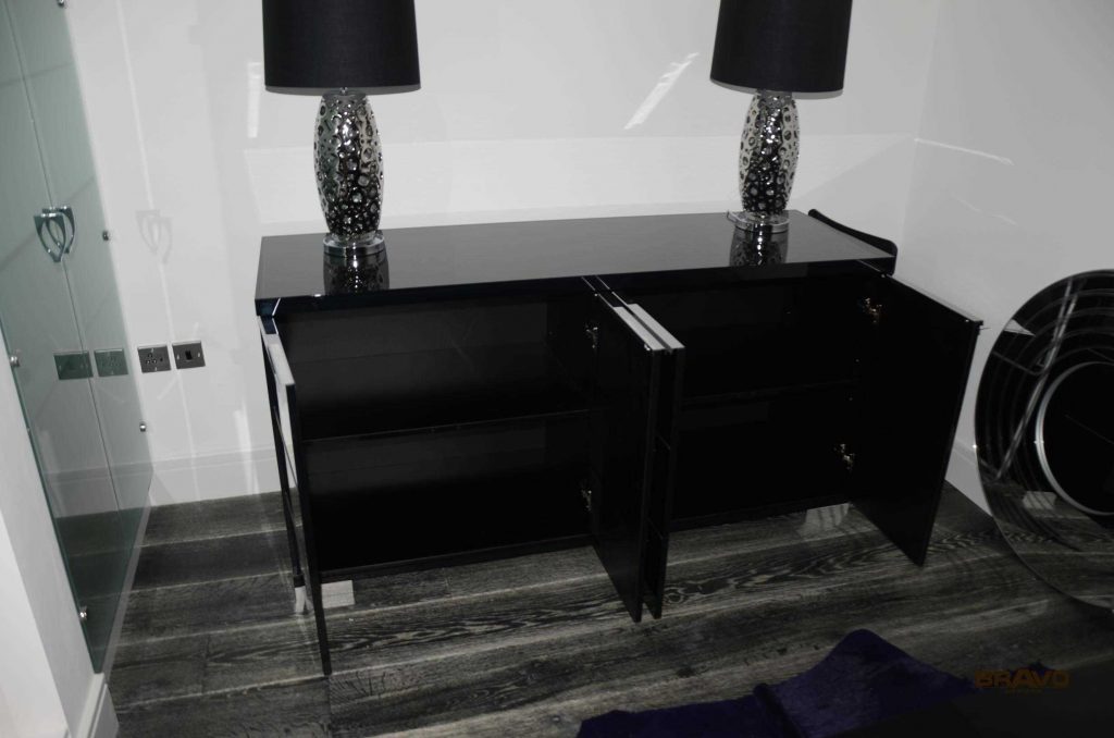 A sleek black console table flanked by two matching lamps with patterned shades, located in a modern room with gray flooring and a glass partition visible to the side, complementing the adjacent fitted wardrobes.
