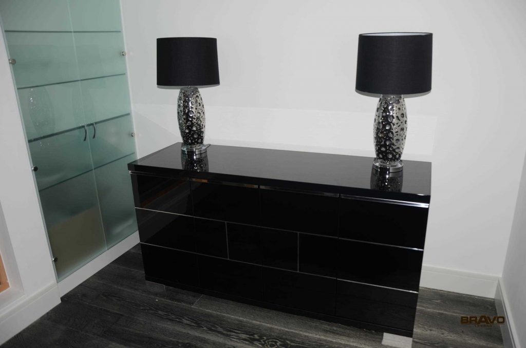 A modern, glossy black dresser with six drawers, located in a room with light gray flooring. On top of the dresser are two identical lamps with black shades and mosaic-style bases beside bespoke furniture.