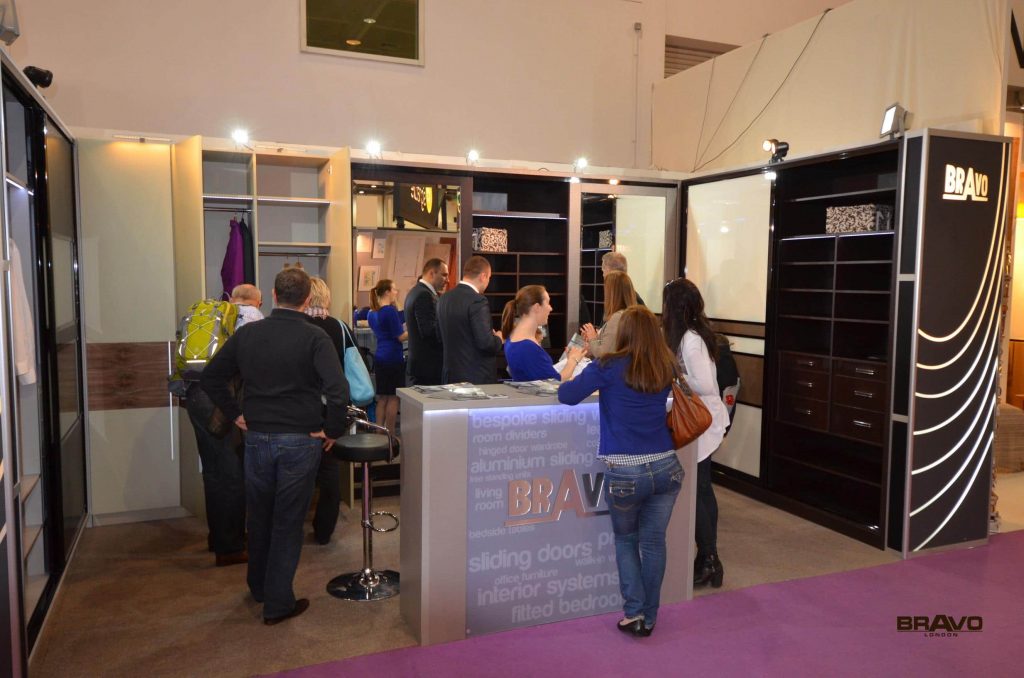 People browsing a modern furniture exhibition with various sliding door designs and bespoke furniture displayed, including several attendees speaking with company representatives.