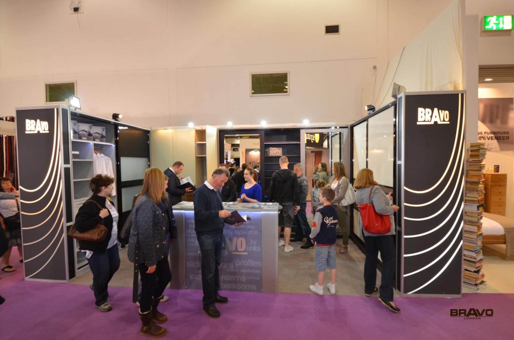 Visitors interacting with exhibitors at a busy trade show booth featuring sleek, sliding door wardrobes and branding.