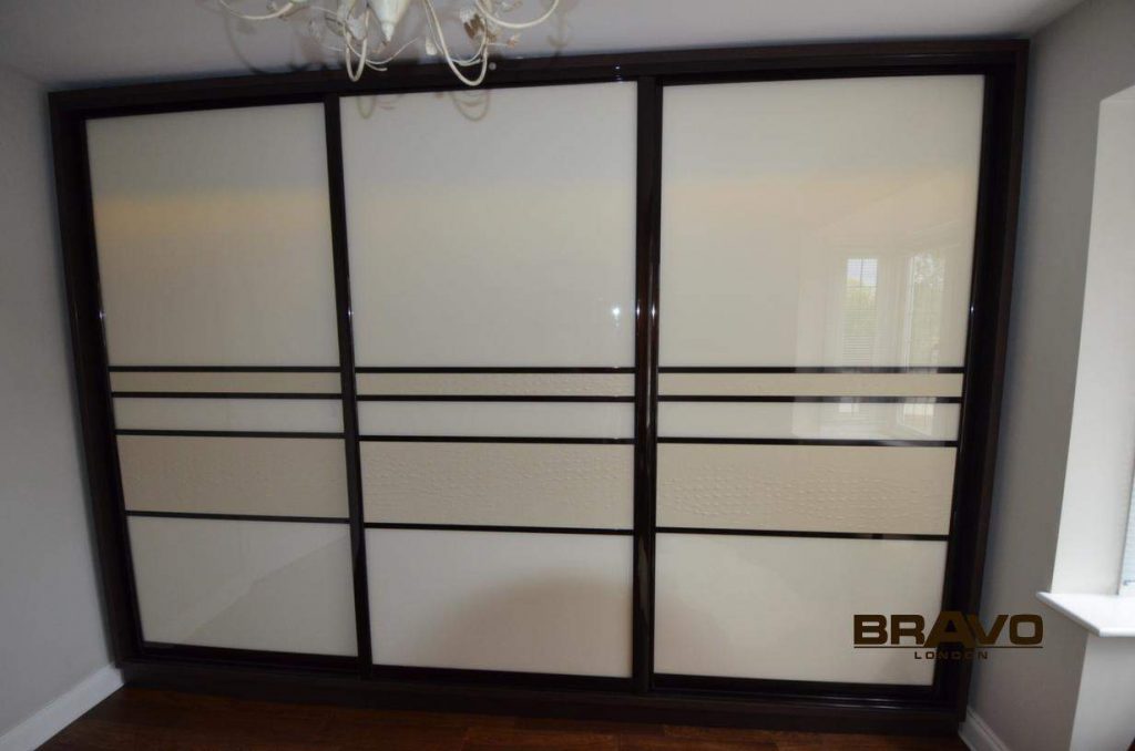 A large, modern bespoke sliding door wardrobe with a black frame and frosted glass panels, featuring horizontal black lines and located in a room with a chandelier and wooden floor.