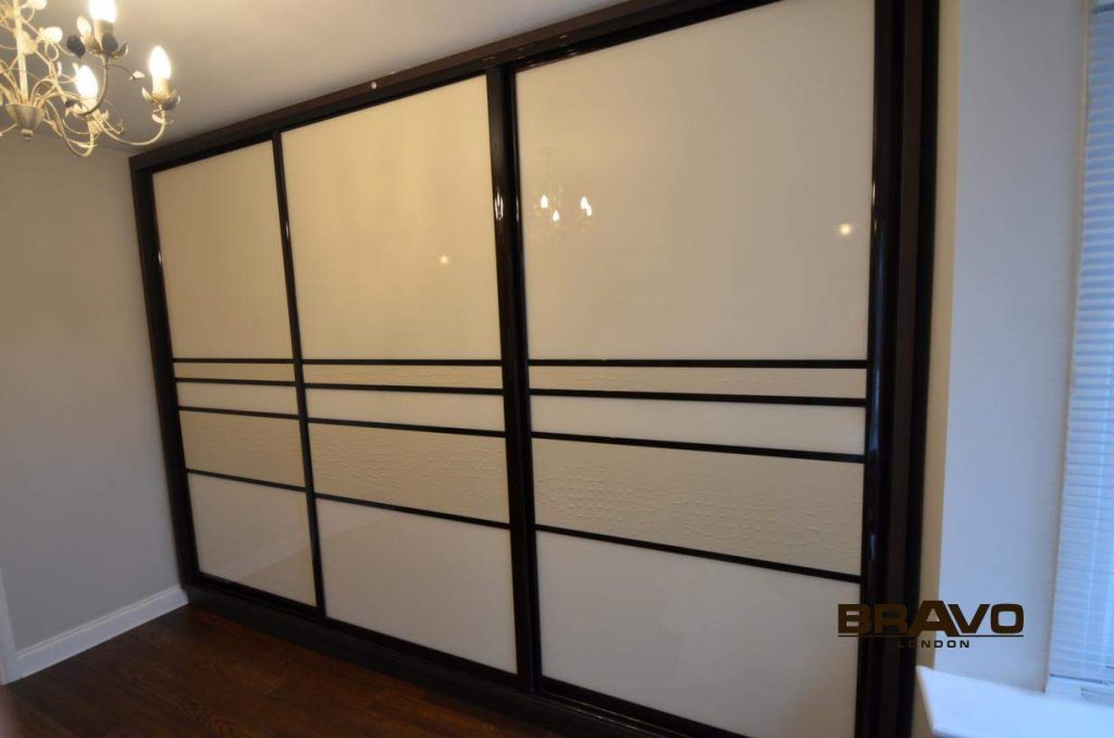 A modern black framed sliding door wardrobe with three large, white opaque panels, installed in a room with wooden flooring and a chandelier visible.