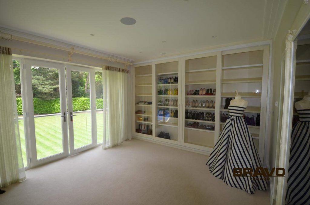 A spacious, well-lit room with large windows and patio doors, featuring a built-in white shelving unit filled with various shoes and two striped mannequins alongside elegant fitted wardrobes.