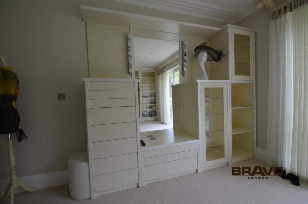 A well-lit modern bedroom featuring a large, white fitted wardrobe with ample storage options, including drawers, shelves, and a center dressing table with a mirror.