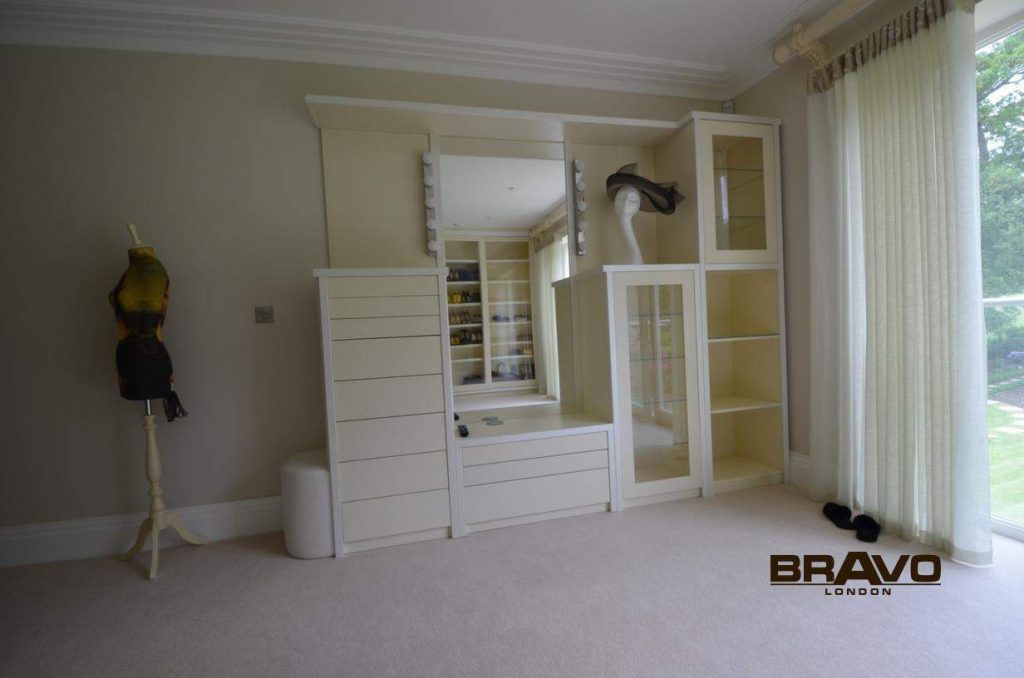 A spacious room featuring a custom-built white storage unit with cabinets, drawers, and shelves, a mannequin with a colorful top, and a large window with curtains. The tailor-made storage includes bespoke furniture that adds both function and style to the space.