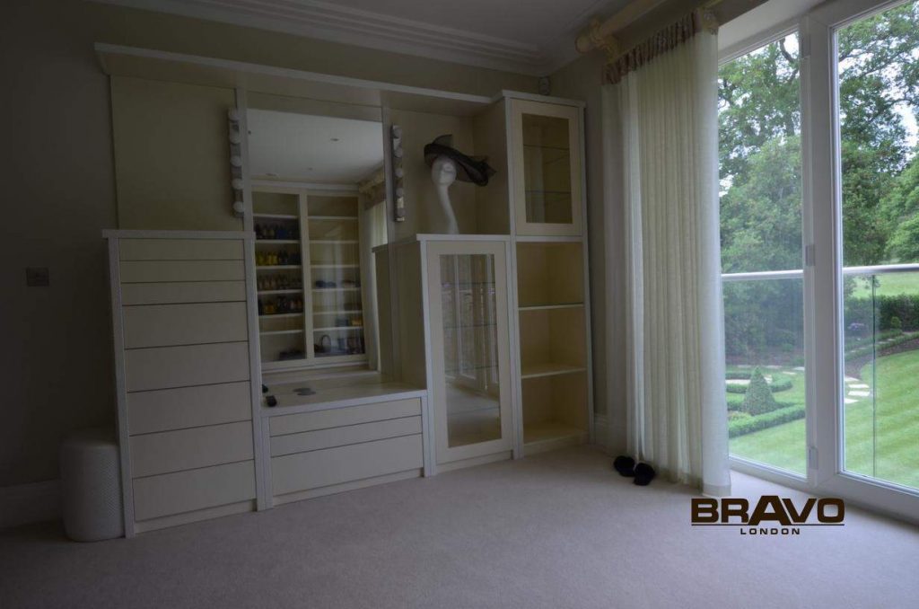An elegant room featuring bespoke white cabinetry along one wall, with shelves, drawers, and a glass-fronted cabinet. The room has a large window, carpeted floors, and a green outdoor view.
