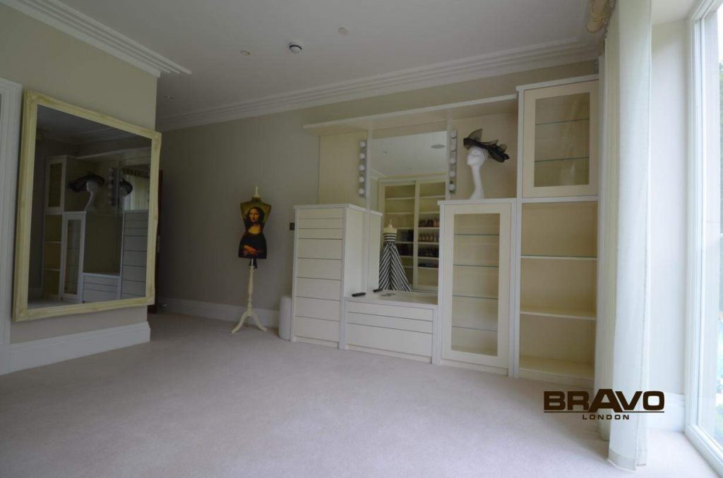 Spacious room with beige carpet featuring a built-in white wardrobe with sliding doors and shelving unit, and a large mirror on the left. A child is partially visible in the background.