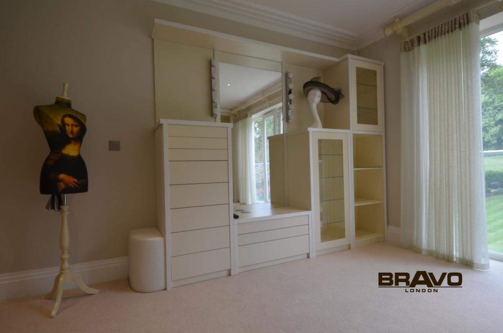 Elegant room featuring a built-in wardrobe with white cabinets and glass doors, a minimalist desk area, and a mannequin displaying a mona lisa print top next to a window overlooking a garden. This fitted bedroom integrates seamlessly with the overall design theme.