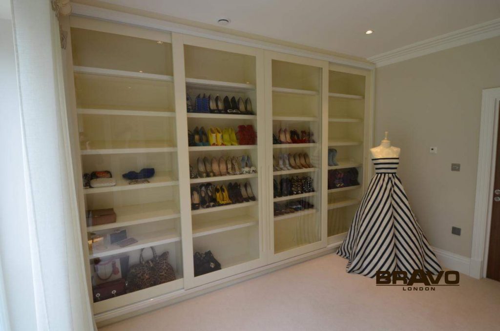 A spacious walk-in closet featuring a large white bespoke furniture wardrobe filled with neatly organized shoes and handbags, and a mannequin displaying a black and white striped dress.
