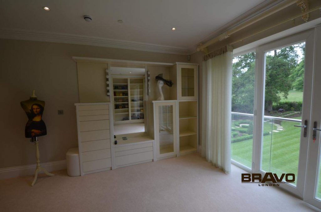 A spacious, modern room with cream carpet, white fitted bedrooms and shelves, large glass doors opening to a green garden, and a portrait on a stand to the left.