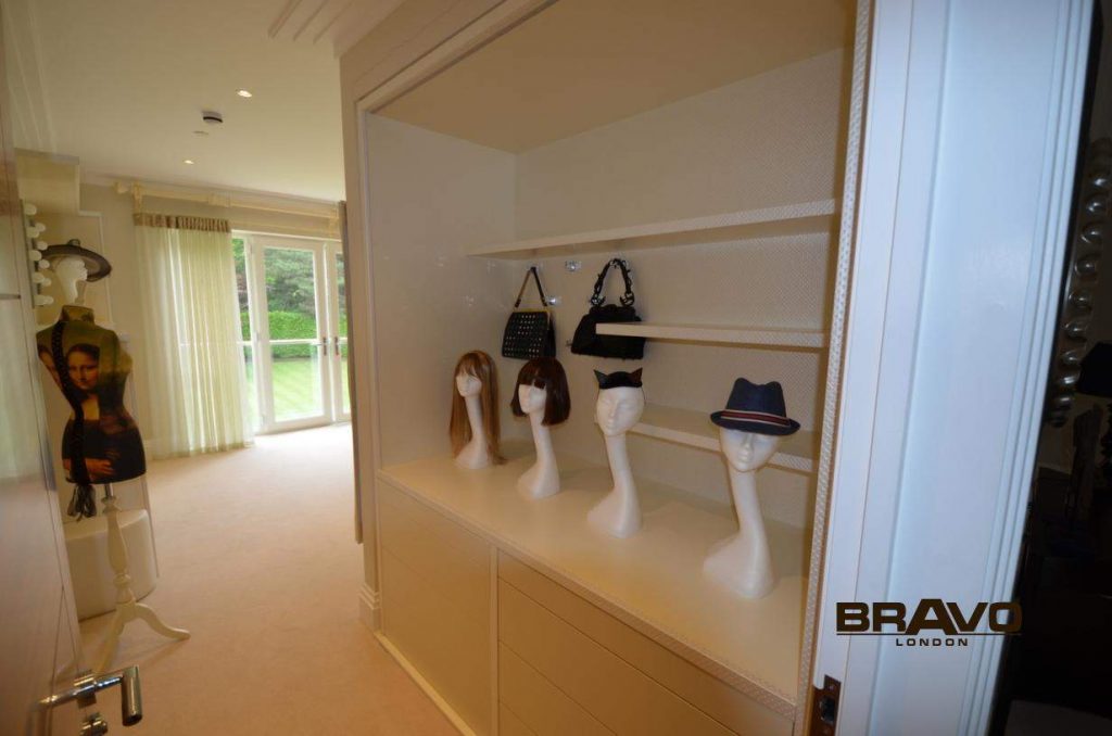A modern walk-in wardrobe room with a display case featuring several white mannequin heads adorned with different wigs and hats. A reflection of a person is subtly visible on the glass door to the left.