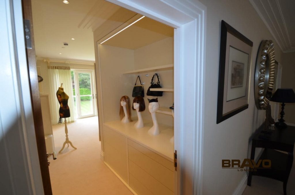A spacious, well-lit dressing area with bespoke white cabinetry and an open wardrobe displaying hanging clothes and accessories. A large mirror and mannequin legs add unique decorative elements.