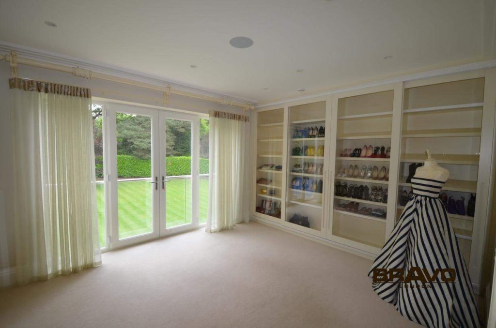 Spacious room with large windows and garden view, featuring a sliding door wardrobe filled with shoes, and a dress on a mannequin. Light curtains complement the soft, neutral decor.