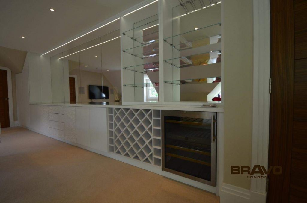 A modern, well-lit room featuring a large white bespoke furniture system with shelves, a wine rack, and glass doors. A sleek TV is also visible. The floor is covered with a light carpet, and wooden sliding door wardrobes are on the left.