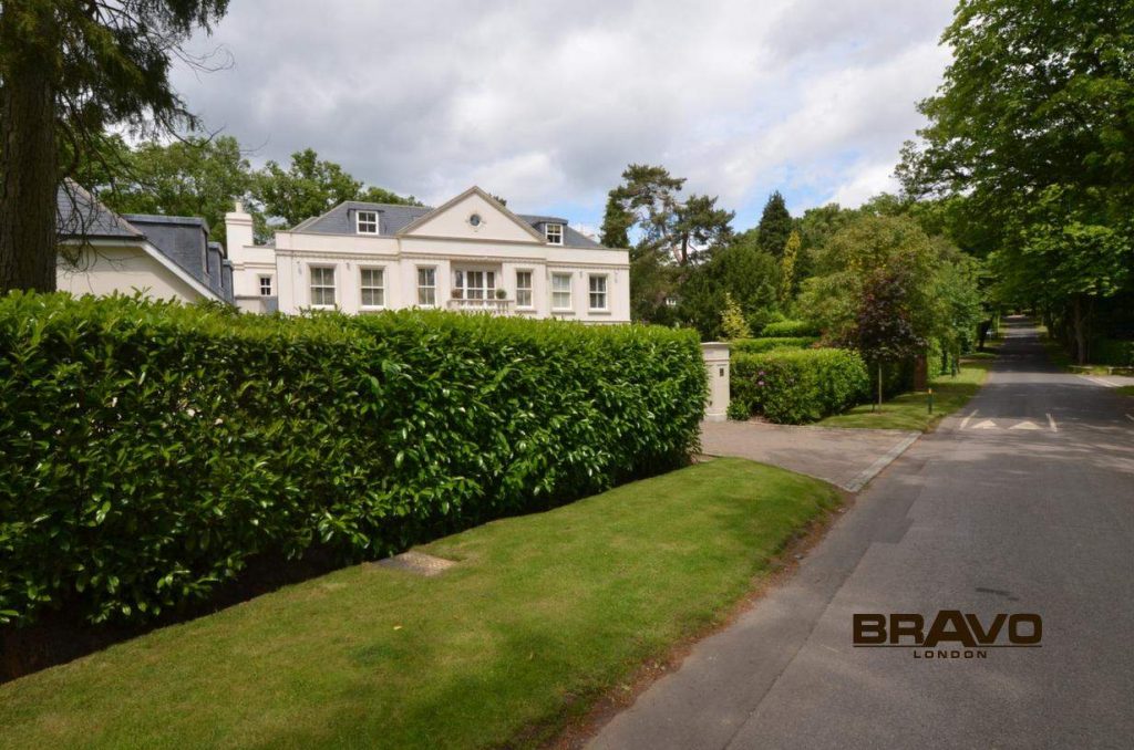 A picturesque suburban house with an elegant, white facade behind lush green hedges, flanked by a paved driveway and surrounded by mature trees. The fitted wardrobes from "Bravo London" are visible in the corner.