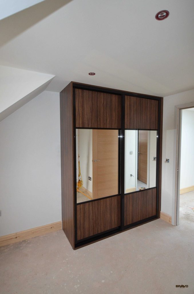 A modern, dark wood fitted wardrobe with mirrored doors installed in a room with unfinished walls and visible construction elements.