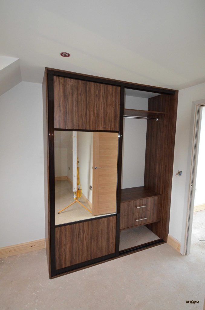 A modern wooden wardrobe with sliding doors, one of which has a mirror. This bespoke furniture piece is in a room with white walls and a light-coloured floor.